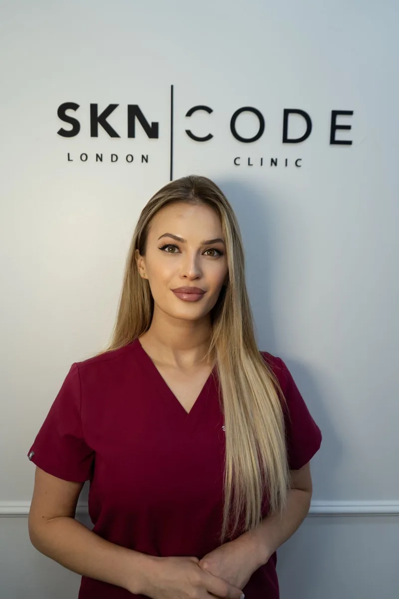 Alkelida, aesthetic practitioner, in burgundy scrubs in front of the SKNCode London Clinic sign